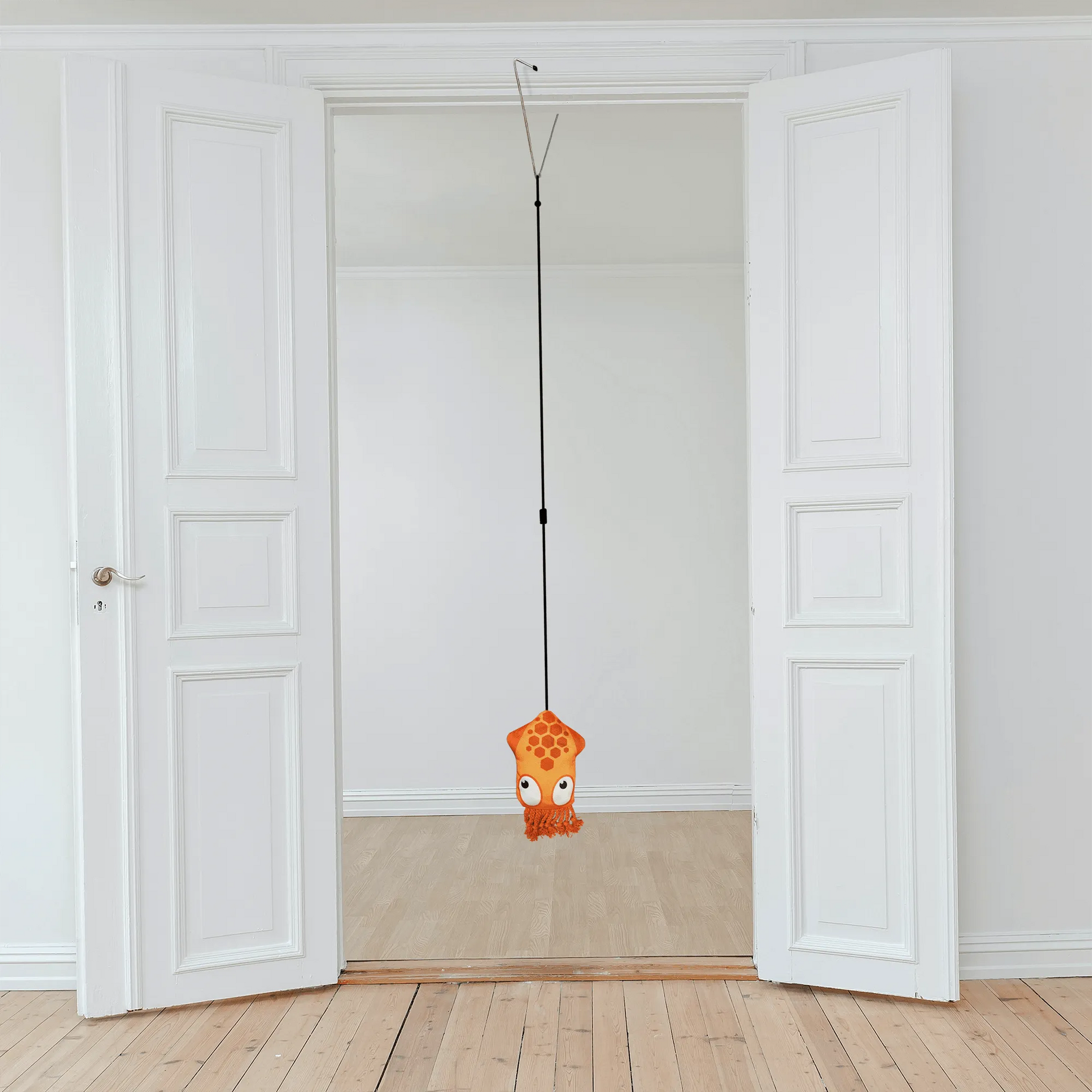 An orange squid catnip toy hangs from a door jamb clip in a bright, empty room with white doors and wood flooring, ready for cat playtime.