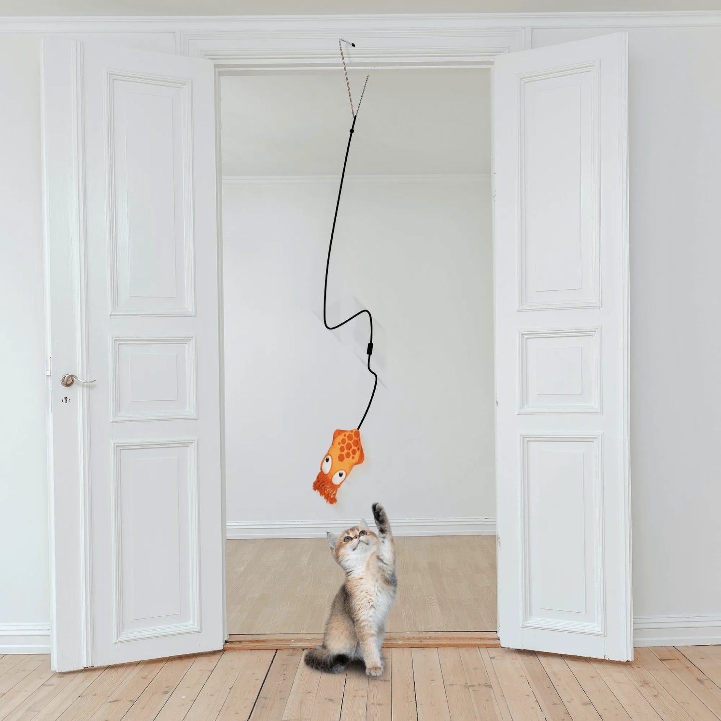 A cat reaches up to bat at an orange squid catnip toy hanging from a door jamb clip in a minimalist room with white doors and wood flooring.